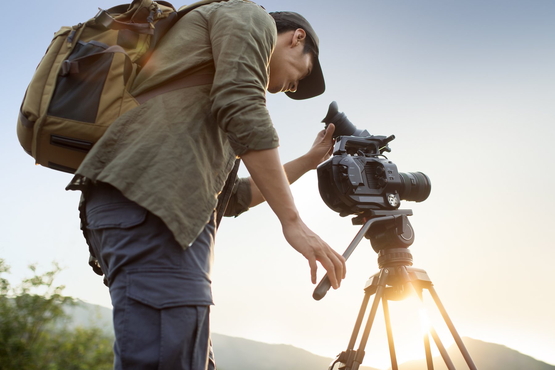 A camera operator with a field pack adjusts a cinema camera on a tripod at golden hour