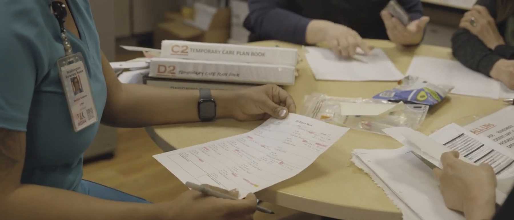 A nurse in teal scrubs holds a hand-marked shift report at a table beside two white binders labeled Temporary Care Plan Book, while colleagues work through paperwork across the table