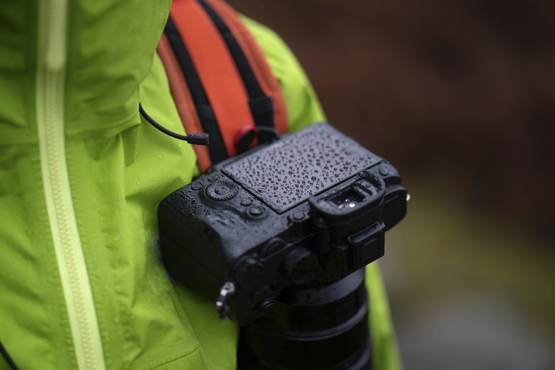 A black mirrorless camera beaded with raindrops, hanging from an orange pack strap against a green rain jacket